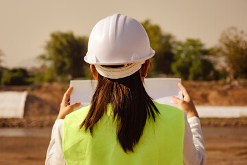 Asian female engineer wearing a helmet and safety vest works and looks at the blueprints for construction plan and design details of the dam.