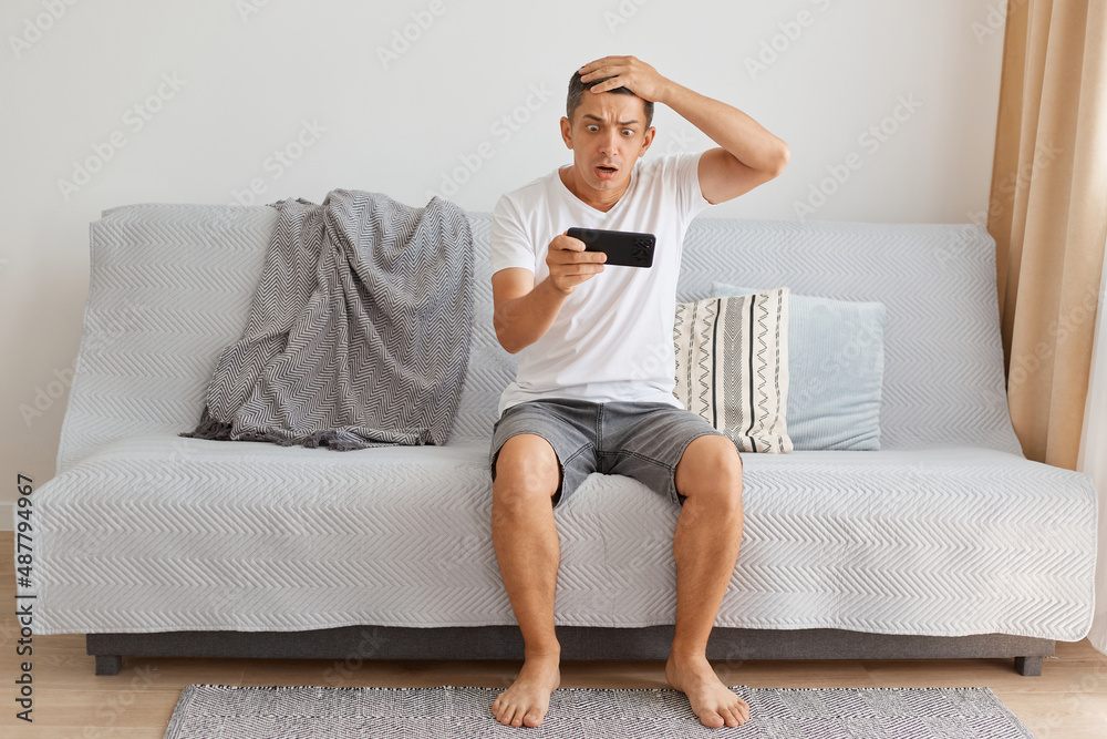 Wall mural Full length of shocked Caucasian dark haired man wearing casual style white t shirt sitting on cough, playing video game, being astonished of level result, keeping hand on head.