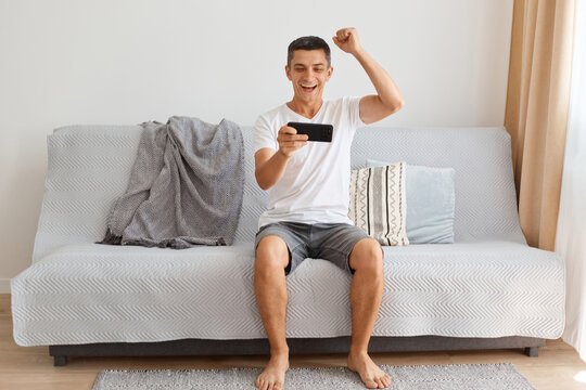 Full length portrait of happy excited smiling man wearing casual style white t shirt sitting on sofa, completed level, clenched fist, celebrating victory, expressing happiness. - Powered by Adobe