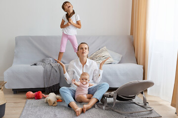 Full length of portrait of depressed mother wearing white shirt and jeans, sitting on floor near sofa and feels desperate, raised arm, don't know what to with screaming children.
