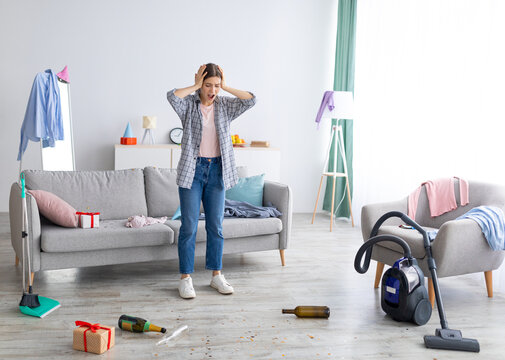 Shocked young woman looking at messy apartment after party, holding head in terror, not knowing where to start cleaning