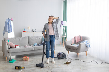 Tired young woman vacuuming her house after party, wiping her forehead, feeling tired of cleaning, free space