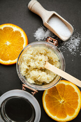 Homemade sugar body scrub in glass jar, decorated with fresh orange slices and wooden spoon with sugar powder on black stone cutting board. Body skin care concept.