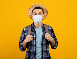 Traveling during coronavirus. Young guy with backpack wearing face mask and straw hat on orange studio background