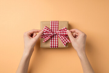 First person top view photo of valentine's day decorations girl's hands tying checkered ribbon bow on craft paper giftbox on isolated beige background