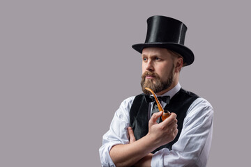 Mature gentleman in black top hat and suit smoking pipe over grey studio background. Rertro styles...