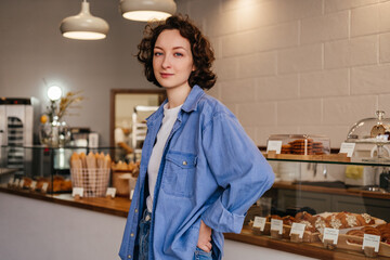 Smiling young woman bakery shop owner standing in front of showcase with pastry products. Small business concept