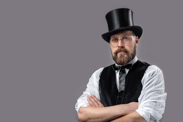 Portrait of confident bearded magician wearing eyeglasses, bow tie and top hat posing in studio...