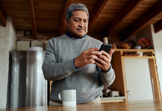 Low angle of healthy multi-cultural elderly male typing a message on smartphone while standing in modern kitchen.