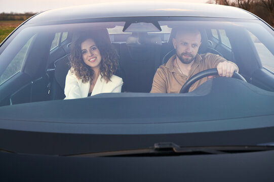 Front View In Black Car Of Focused Male Driver And Smiling Female Passenger Traveling By Car Out The Town.