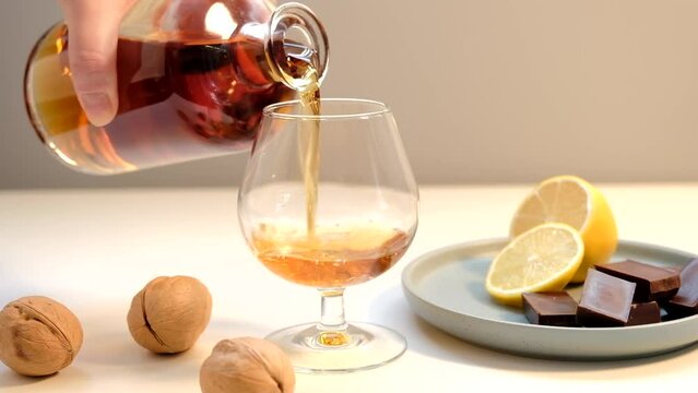 The hand pours cognac from the bottle into the wine glass. Next to it on a plate is a lemon and pieces of chocolate.