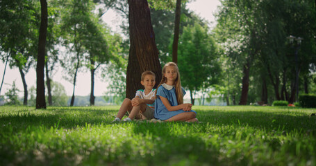 Smiling siblings play on grass under tree. Happy children sitting on lawn.