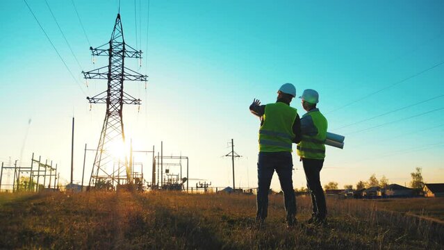 Teamwork industry a concept. Two engineers electric experts in protective helmets discussing the construction of a power line. Silhouette Of Engineers Standing On Field With Electricity Towers.