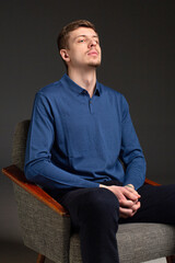 Attractive young blond european man with short beard and mustache isolated on black sitting in armchair