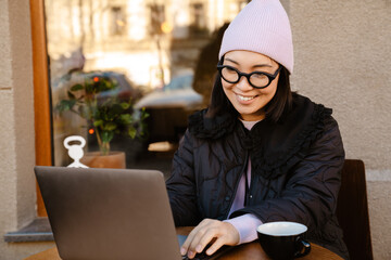 Young asian woman smiling and typing on laptop at cafe outdoors