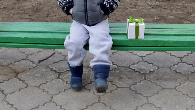 little kid is sitting on a green park bench with a gift box in hand or on the seat. white box with green ribbon. present to his beloved girl. waiting the girl friend. boy is alone outside. march 8, 
