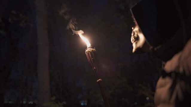 A disguised man in a skeleton or death mask holds a tiki torch in his hand and gazes fascinated at the fire at a Halloween party at night. Bamboo torches. Unrecognizable person. Stick burns fire.