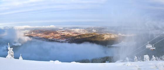 The fog rolls through the mountain valley, at the foot of the mountains there is a small town of Stronie Slaskie, a view from the snow-capped Czarna Gora mountain on a winter day.