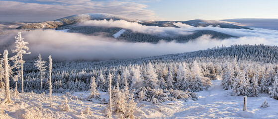 Through the valleys, the fog climbs the snow-capped mountain range of the Snieznik Massif just before sunset, the view from the top of Czarna Gora in winter.