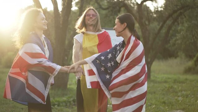Young international business partners stacking hands in slow motion smiling standing in sunrays in summer spring park. Happy confident man and women celebrating success outdoors