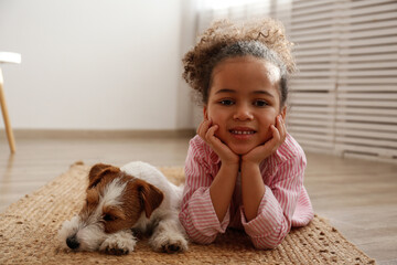 Little black girl playing with her friend, the adorable wire haired Jack Russel terrier puppy at home. Preschooler with rough coated pup lying on the floor. Interior background, close up, copy space.