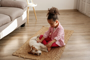 Little black girl playing with her friend, the adorable wire haired Jack Russel terrier puppy at home. Preschooler with rough coated pup sitting on the floor. Interior background, close up, copy space
