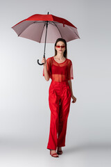 full length of young woman in red trendy outfit and sunglasses standing under umbrella on grey.