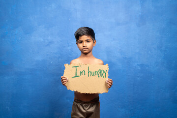 Sad Indian poor kid holding I am hungry sign board by looking at camera on blu background with copy...