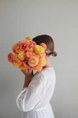 Conceptual portrait of young woman wearing white cotton dress covering her face with lush ranunculus. Female hiding behind beautiful bouquet of bright flowers. Close up, copy space, white background