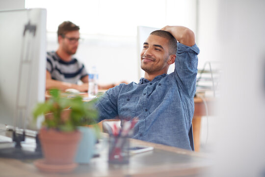 He Knows He Has The Answer. Young Creative Professional Smiling At His Pc In A Bright Communal Office Space.