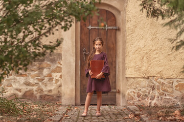 Fototapeta na wymiar Portrait of a beautiful little girl with an old encyclopedia in her hands in the courtyard of the house.