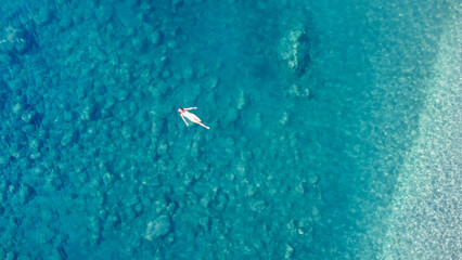 Young pretty woman in a white one piece swimsuit svimming on back in turquoise sea water. Woman lies on the water transparent water. Aeriale drone photo