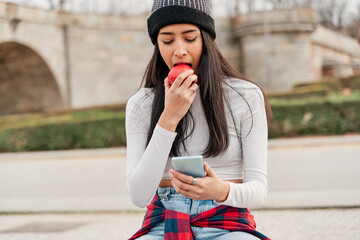 Fototapeta premium Funny hungry girl with an appetite bites into a red apple sitting in the park looking at her smart phone. healthy life concept