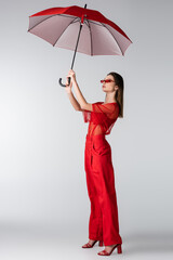 full length of young model in red trendy outfit and sunglasses holding umbrella above head on grey.