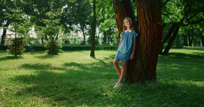 Dreamy girl lean tree trunk in park. Smiling kid observing nature around