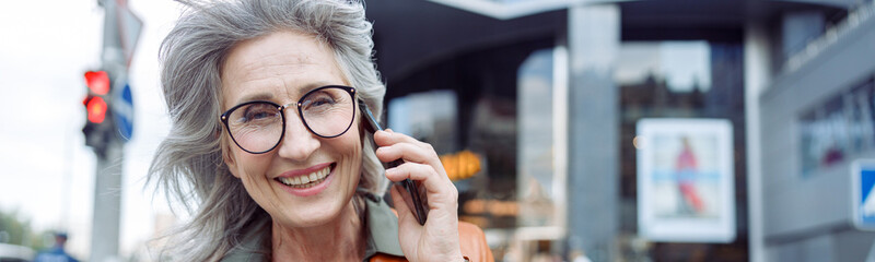 Happy grey haired senior woman with glasses in leather jacket talks on mobile phone on modern city...