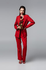 full length of young woman in trendy red suit holding rose while standing with hand on hip on dark grey.