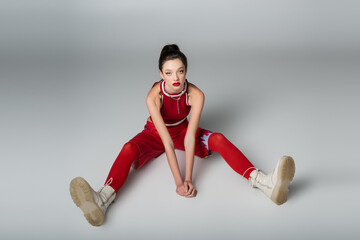 full length of stylish young woman in red sportive outfit and boots sitting on grey.
