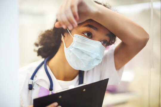 Young Tired Overworked African American Medical Worker Wearing Protective Face Mask With X-ray Scan In Hands Looking Out Window With Frustrated Expression. Healthcare Workers During COVID-19 Pandemic