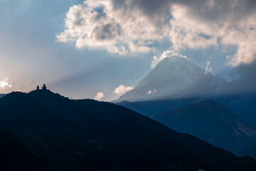 Distant view on Gergeti Trinity Church in Stepansminda, Georgia. The church is located the Greater Caucasian Mountain Range.Clouds are covering the snow-capped Mount Kazbegi in the back.Sunrise,sunset