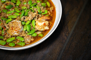 Close up Thai Stir-Fried Stink Beans with Shrimps and spicy shrimp paste sauce in white dish bowl on dark wood table.