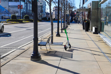a black metal bench and a white and green electric scooter on a sidewalk lined with bare winter...