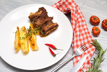 Fried lamb ribs with baked potatoes slices. Thyme herbs. One portion on a big white plate. Table top decorated with checkered cloth, dried tomatoes and rosemary herb.