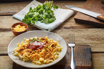 Pasta bolognese garnished with greens and cheese in a plate on a wooden table on a glossy board next to a fork and greens.
