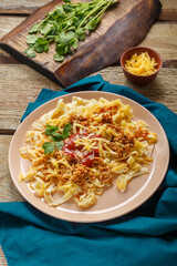 Pasta bolognese garnished with greens and cheese in a plate on a blue napkin on a wooden table next to a fork and greens on a board and grated cheese.