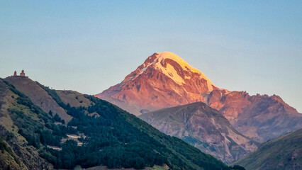 Distant view on Gergeti Trinity Church in Stepansminda, Georgia. The church is located the Greater Caucasian Mountain Range. Clear sky above the snow-capped Mount Kazbegi in the back. Sunrise, sunset