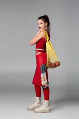 full length of trendy young model in red sportive outfit holding string bag with apples on grey.