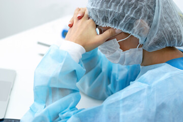 Fototapeta premium A tired female doctor wearing face mask and surgical overalls is sitting at her desk in the medical office with her eyes closed.Healthcare and medical concept.