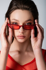 Fototapeta premium young model adjusting red trendy sunglasses isolated on grey.