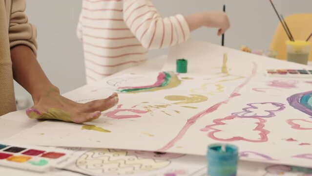 Cropped slowmo of unrecognizable diverse kids having fun painting on big sheet of paper together. Boy making colorful handprints